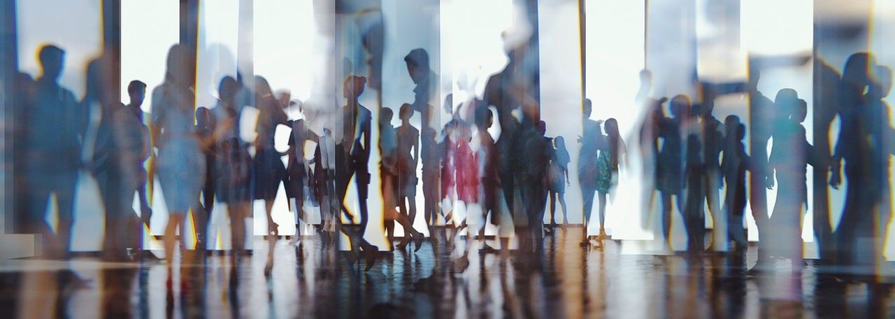 Blurry silhouettes of people walking in an abstract space with light streaming in from windows behind them.